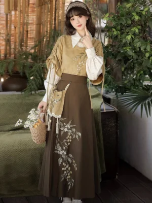 Front three-quarter view of a woman wearing a hazelnut milk tea modern Hanfu blouse and skirt set with embroidered mustard vest, white lantern sleeve shirt, brown pleated skirt, and tassel waist pouch