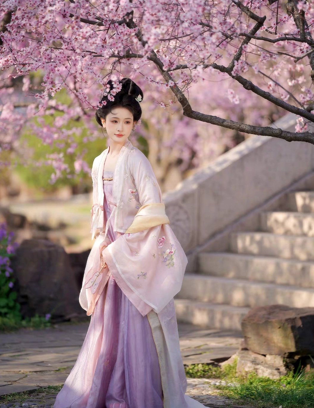 Side view of Tang Dynasty hanfu outfit with floral pink outer robe, gradient yellow cuffs, flowing lavender pleated qixiong skirt and draped shawl in a classical garden