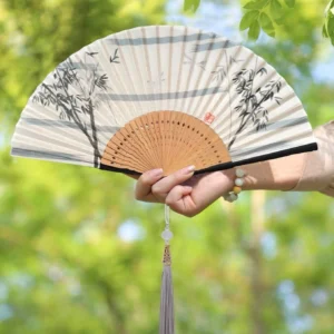Ink bamboo Chinese folding hand fan with carved wooden ribs, gray purple tassel and red seal detail held outdoors