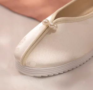 Close-up of ivory traditional Chinese Hanfu flat shoe showing pearl front strap, pale apricot piping, floral texture, and thick white rubber sole