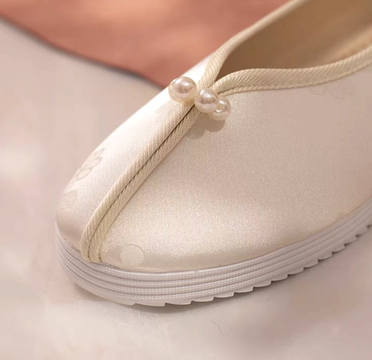 Close-up of ivory traditional Chinese Hanfu flat shoe showing pearl front strap, pale apricot piping, floral texture, and thick white rubber sole