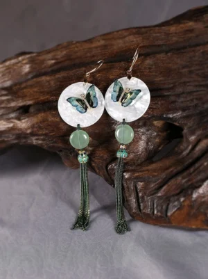 Jade green butterfly tassel Chinese earrings with round moonlike shell pendant displayed on gray fabric beside dark wood decor