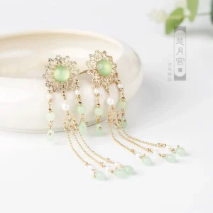Pair of classical Chinese hair pins with gold floral filigree, light green centerpiece stones, pearl accents, and dangling green bead tassels displayed on a white ceramic tray