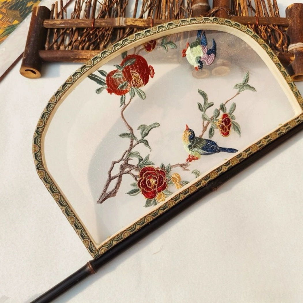 Side oblique view of a knife-shaped Chinese hand fan with a long dark brown bamboo handle, vivid pomegranate and flower embroidery, colorful birds, and traditional geometric brocade edging