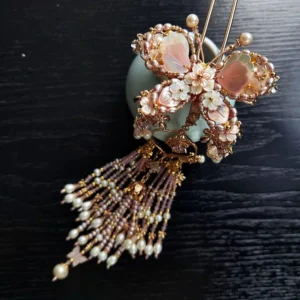 Large butterfly Hanfu hairpin in rose gold and soft pink with pearl accents mother of pearl petals and layered beaded tassels beside a celadon vase