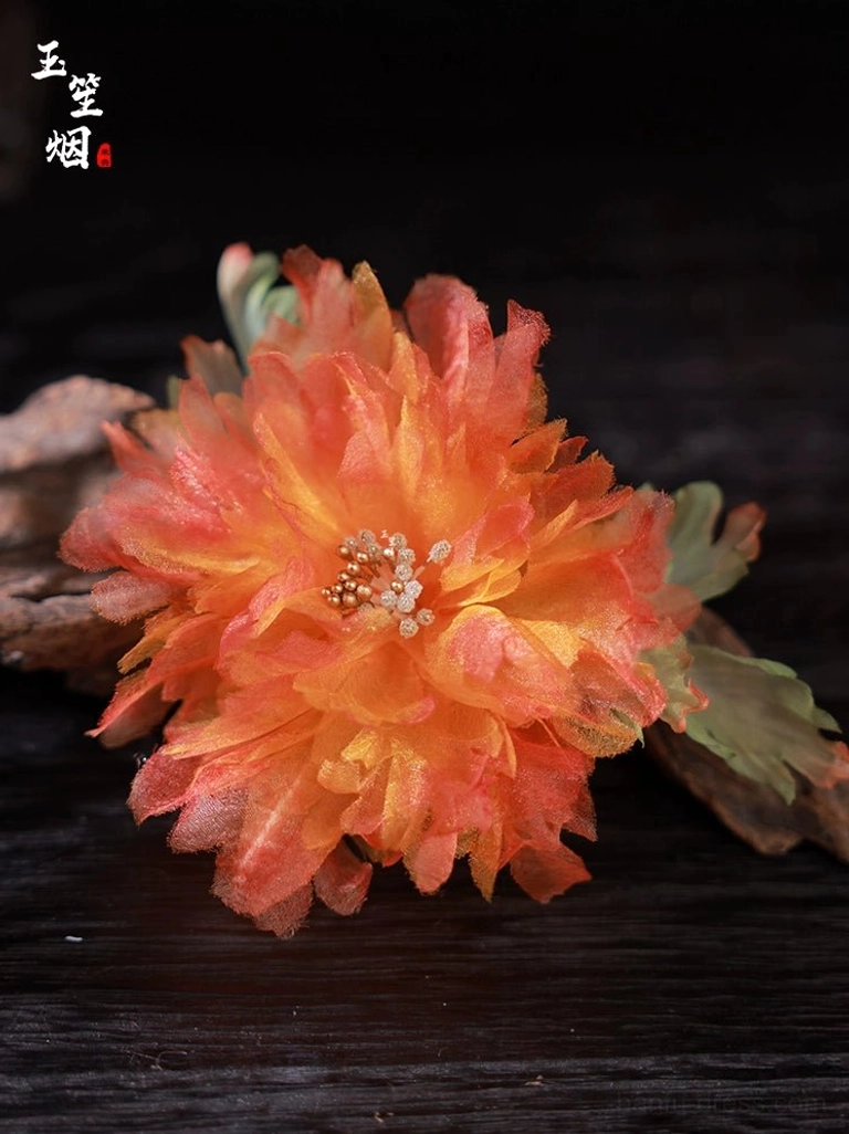 Close-up of a large orange and yellow layered silk peony hairpin with beaded flower center and green foliage on a dark wooden background