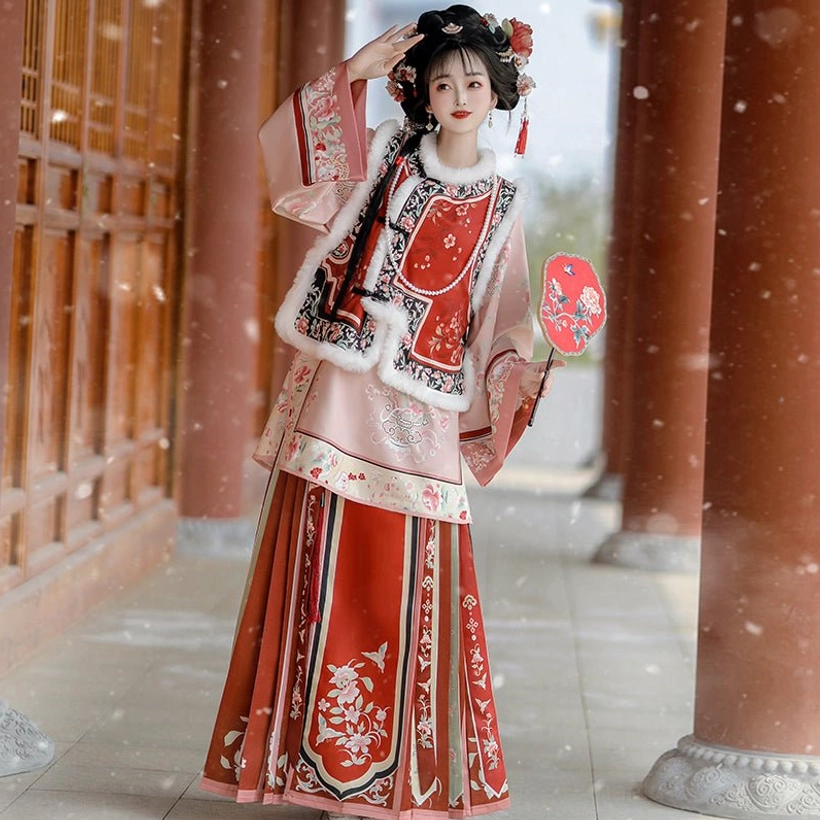 Red Late Qing Han women winter outfit with embroidered floral vest layered wrap skirt and plush collar trim photographed in a snowy wooden corridor