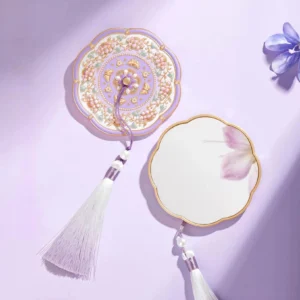 Lavender blossom Chinese pocket mirror with scalloped gold-edged frame, ornate floral back cover, white and purple tassel, faux pearl details, and flower petals on a light purple background