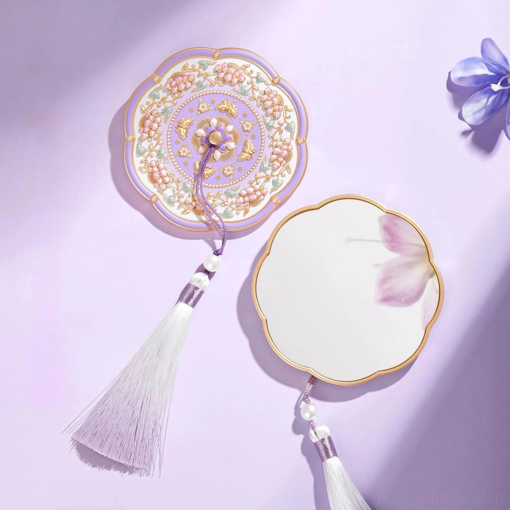 Lavender blossom Chinese pocket mirror with scalloped gold-edged frame, ornate floral back cover, white and purple tassel, faux pearl details, and flower petals on a light purple background