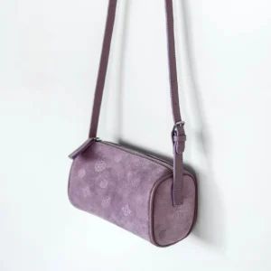 Lavender suede Chinese barrel crossbody bag shown hanging from the side with embossed floral motifs and a long shoulder strap on a white studio background
