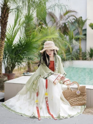 High-angle seated view of a woman wearing a flowing mint green Song Dynasty style Hanfu outer robe with a cream skirt, red and green trim, straw hat, and woven tote bag