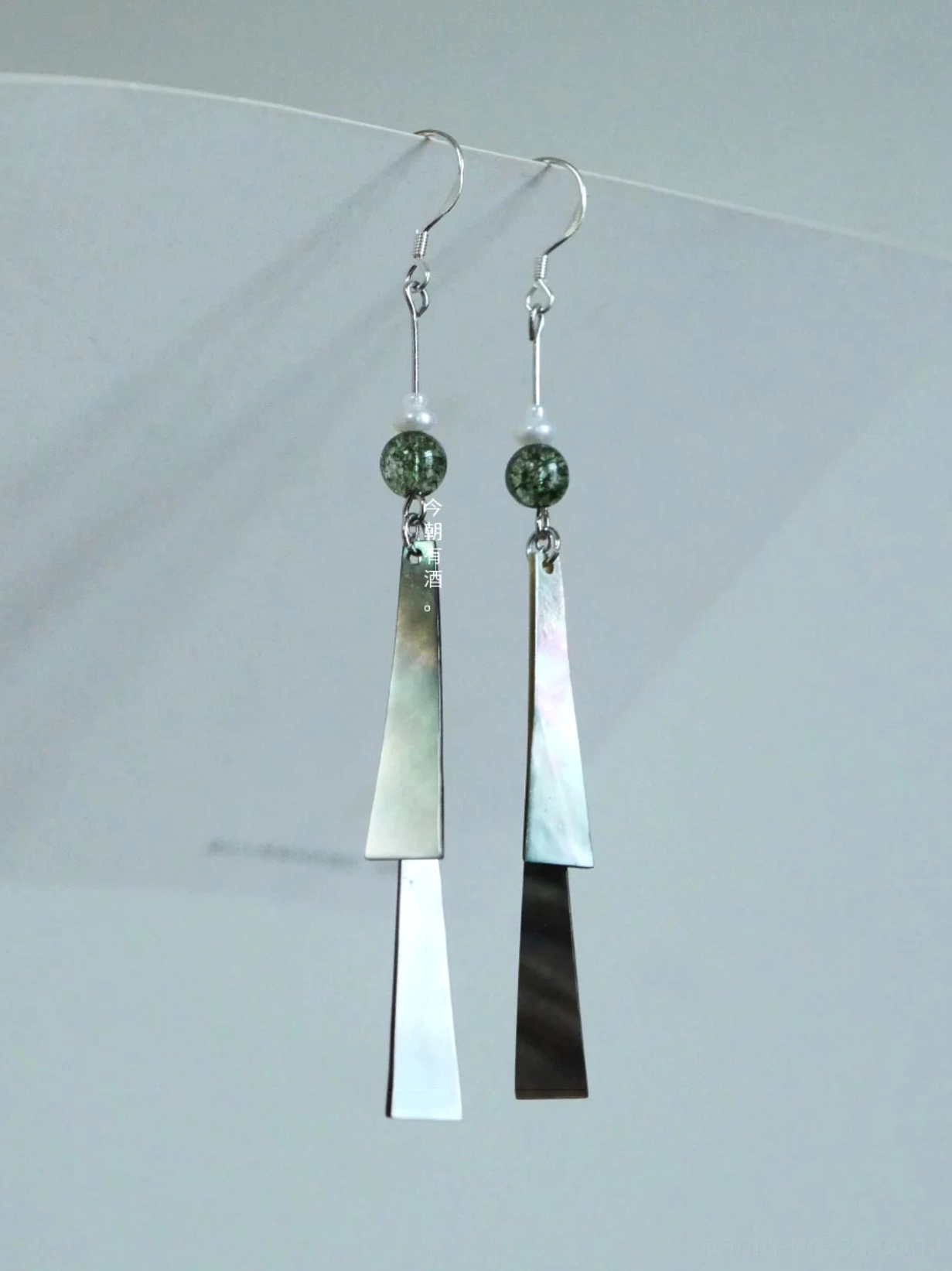 Letter from the Wind green shell tassel Chinese hook earrings displayed on a white board with silver hooks and speckled green beads