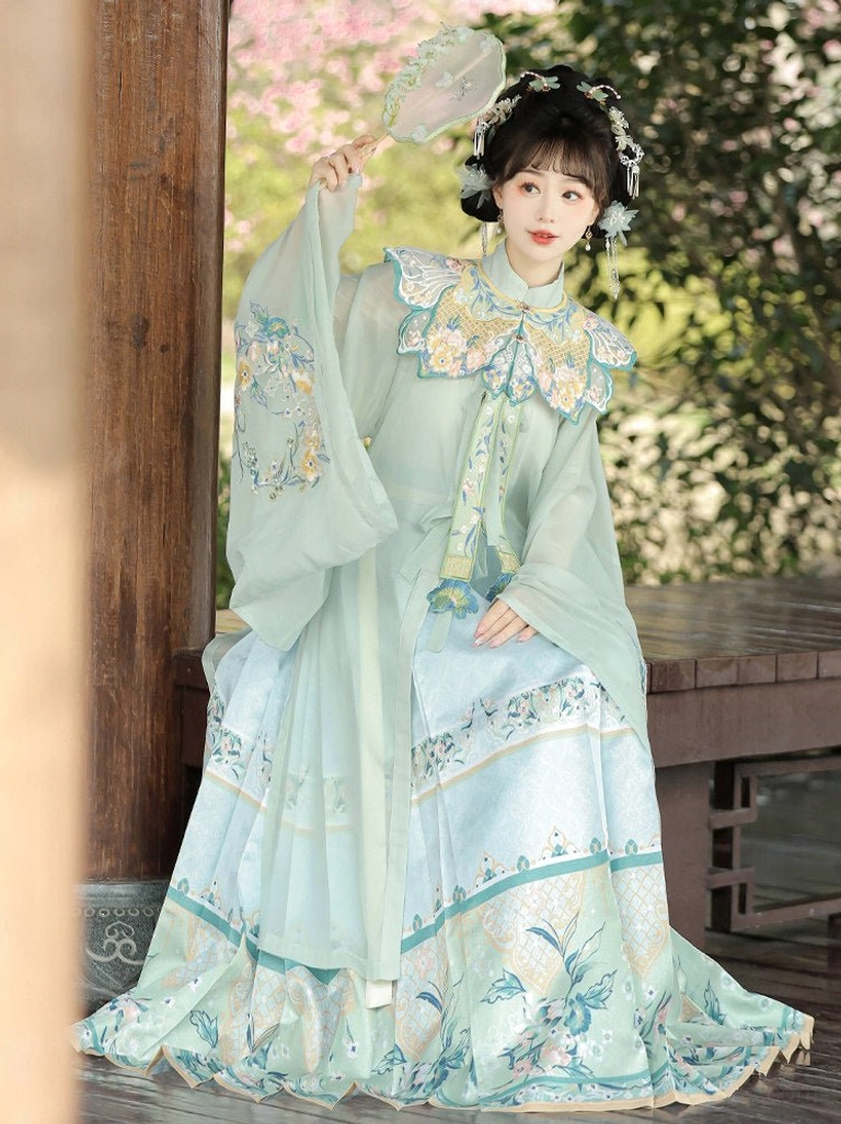 Light cyan traditional Chinese Hanfu dress with embroidered floral and bird cloud collar, flowing robe, mamian skirt, and folding fan in a wooden pavilion