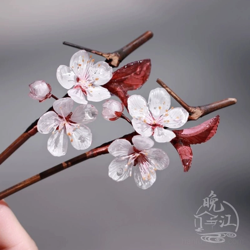 Close-up of Lihua plum blossom Chinese hair stick held by hand, showing white flower petals, pink buds, red leaves, and slender brown hairpin shaft