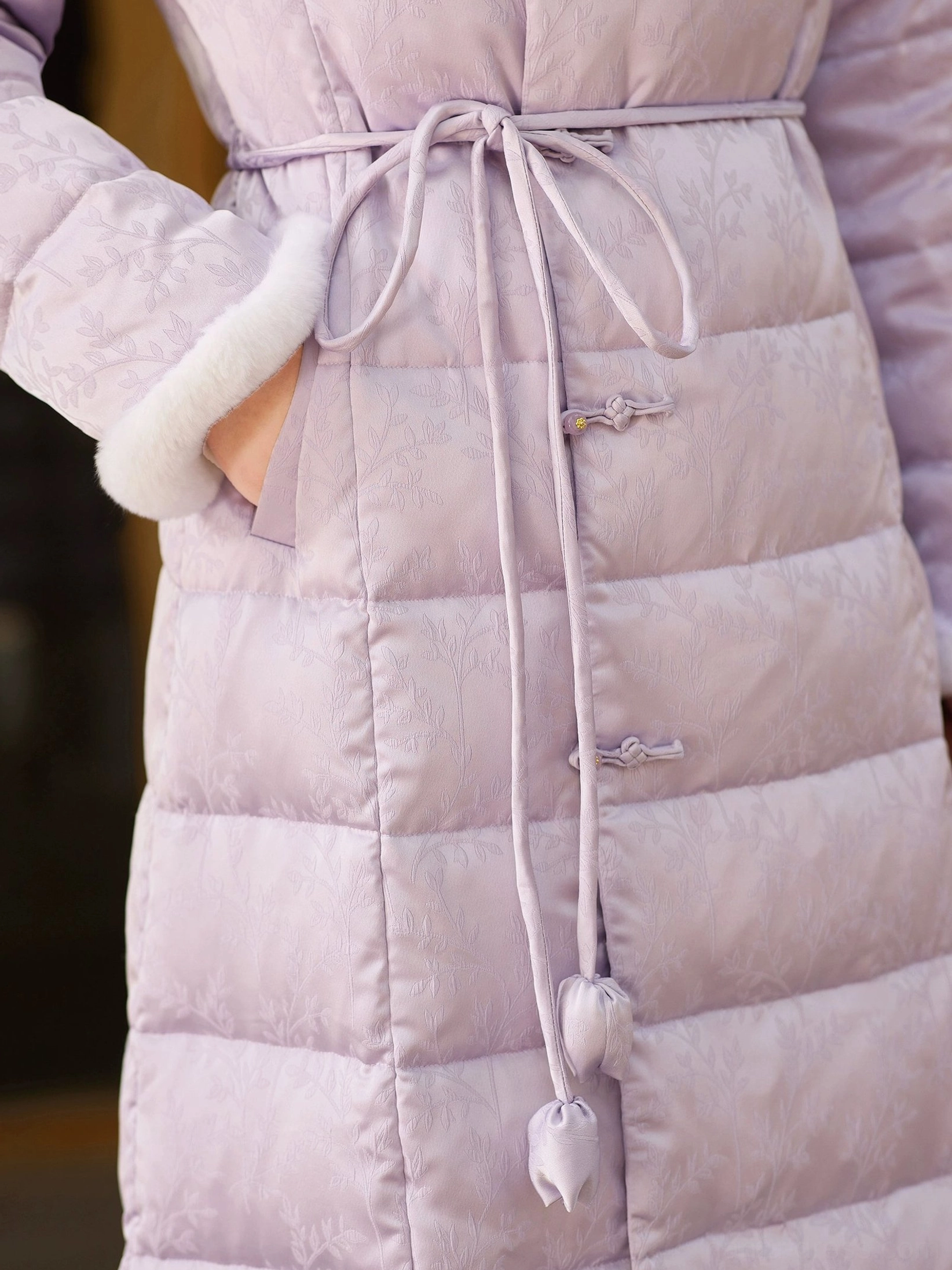 Close-up of a lilac qipao-inspired down coat showing the bow-shaped drawstring, fuzzy cuffs, Chinese frog buttons, quilted fabric, floral lining and side pocket