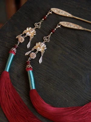 Pair of Liluo Chinese tassel hair sticks with deep red silk fringe, floral pendants, red beads, and teal wrapped details on a dark wooden surface