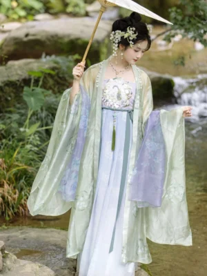 Full body side view of a lily of the valley Tang Dynasty Hanfu dress with sage green sheer wide sleeves, gradient purple blue skirt, embroidered bodice, and waist tassel pendant in a traditional Chinese waterside garden