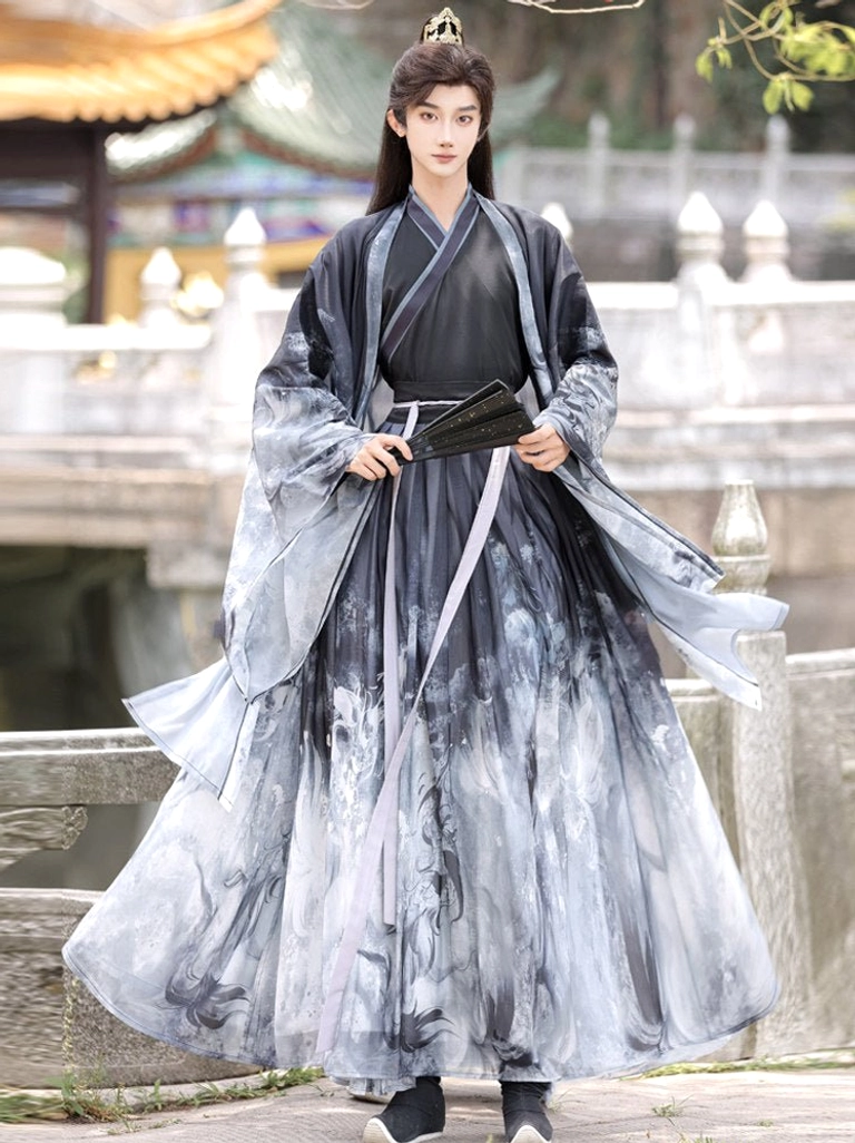 Male model wearing black and grey ink-wash Xianxia hanfu set with dragon print, wide sleeves, lavender waist sash and black folding fan in a traditional Chinese garden