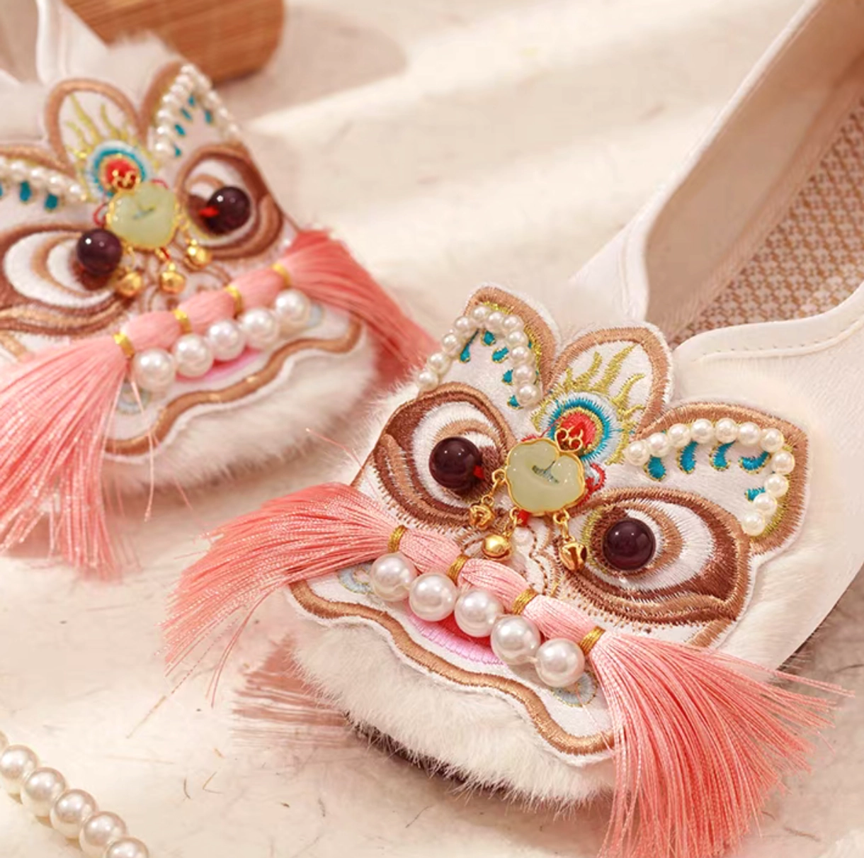 Close-up side view of a traditional Chinese Hanfu shoe with detailed lion head embroidery, brown resin-style eyes, jade-inspired ornament, pink tassels, pearl strands, fuzzy toe trim, and woven insole