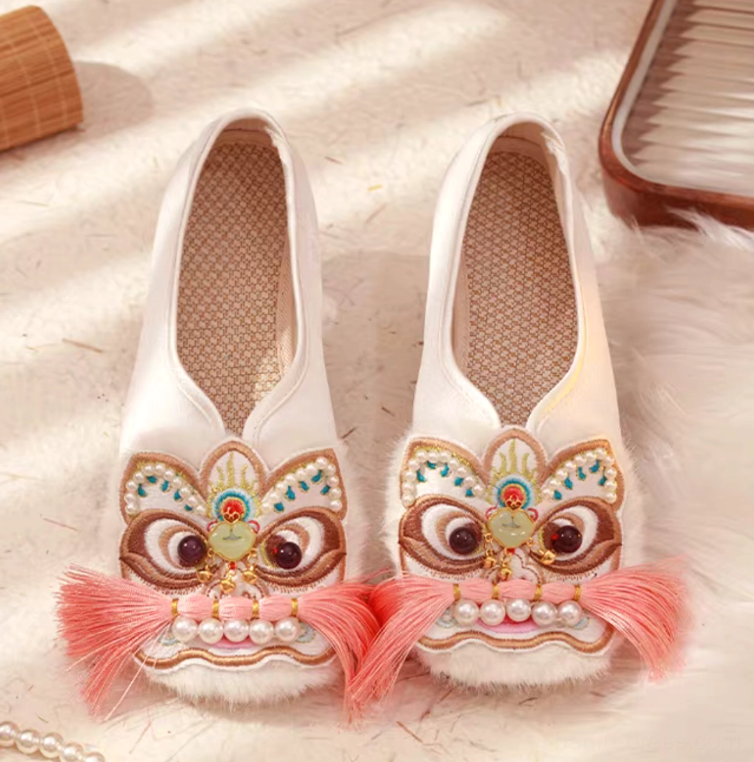 Pair of traditional Chinese lion embroidered Hanfu shoes with off-white canvas uppers, pink tassels, pearl ornaments, fuzzy toe trim, and breathable woven insoles displayed on a decorative surface