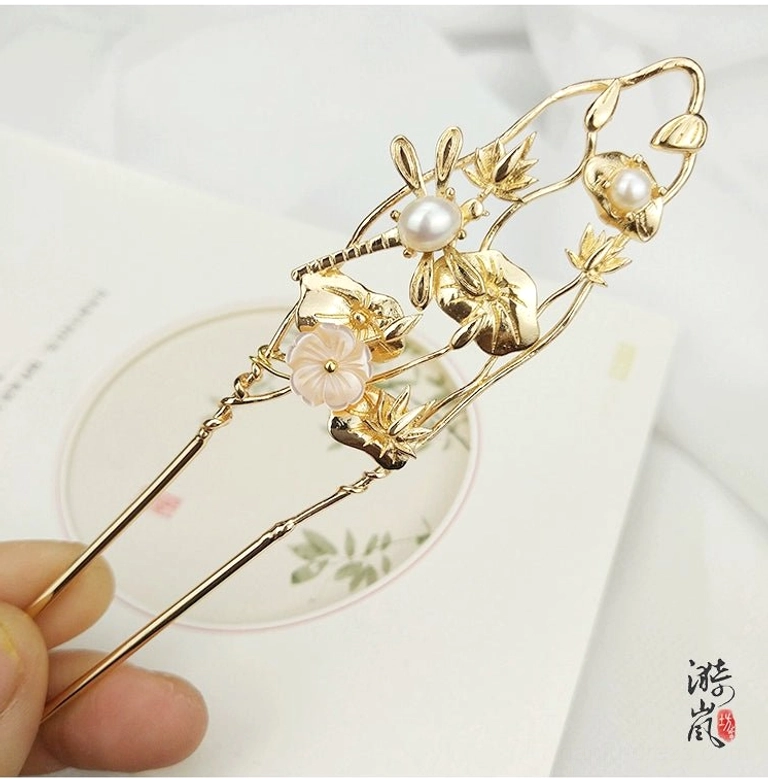 Handheld view of Lixia gold floral double-prong hairpin showing openwork leaf details, faux pearls and pink resin flowers