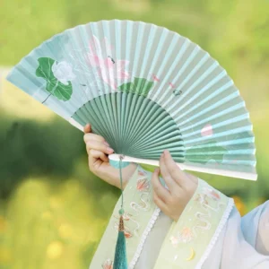 Lotus Chinese folding hand fan with mint green surface, dark green bamboo ribs, and matching tassel held with a Hanfu outfit outdoors