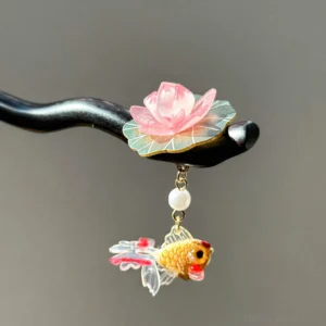 Close-up of a traditional Chinese hair stick with a pink lotus flower, green lotus leaf, white pearl pendant, golden goldfish charm, and curved black wooden pin