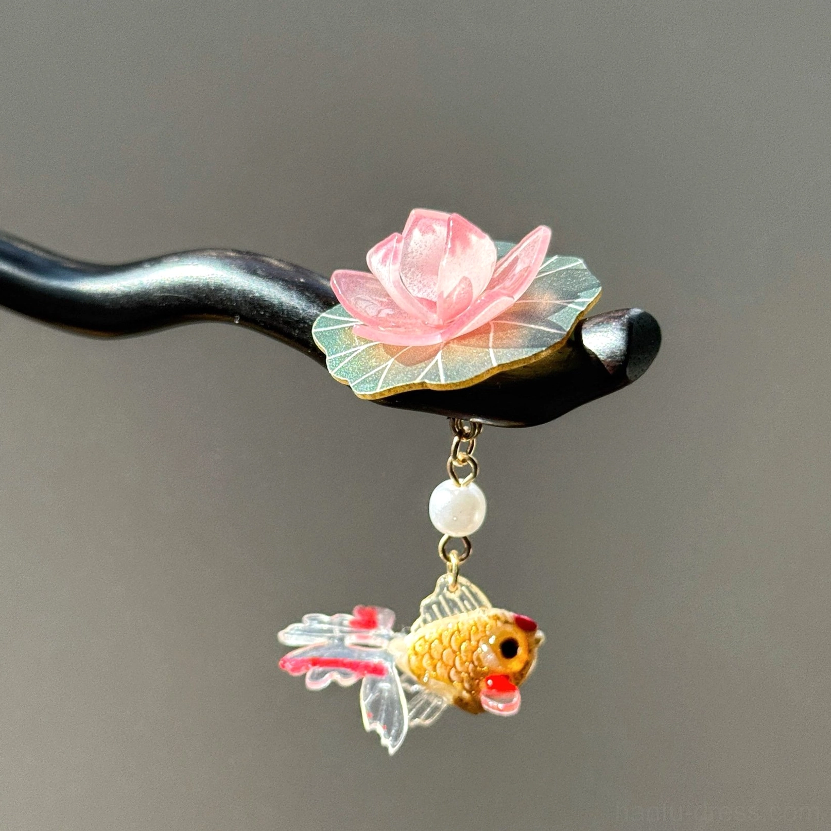 Close-up of a traditional Chinese hair stick with a pink lotus flower, green lotus leaf, white pearl pendant, golden goldfish charm, and curved black wooden pin