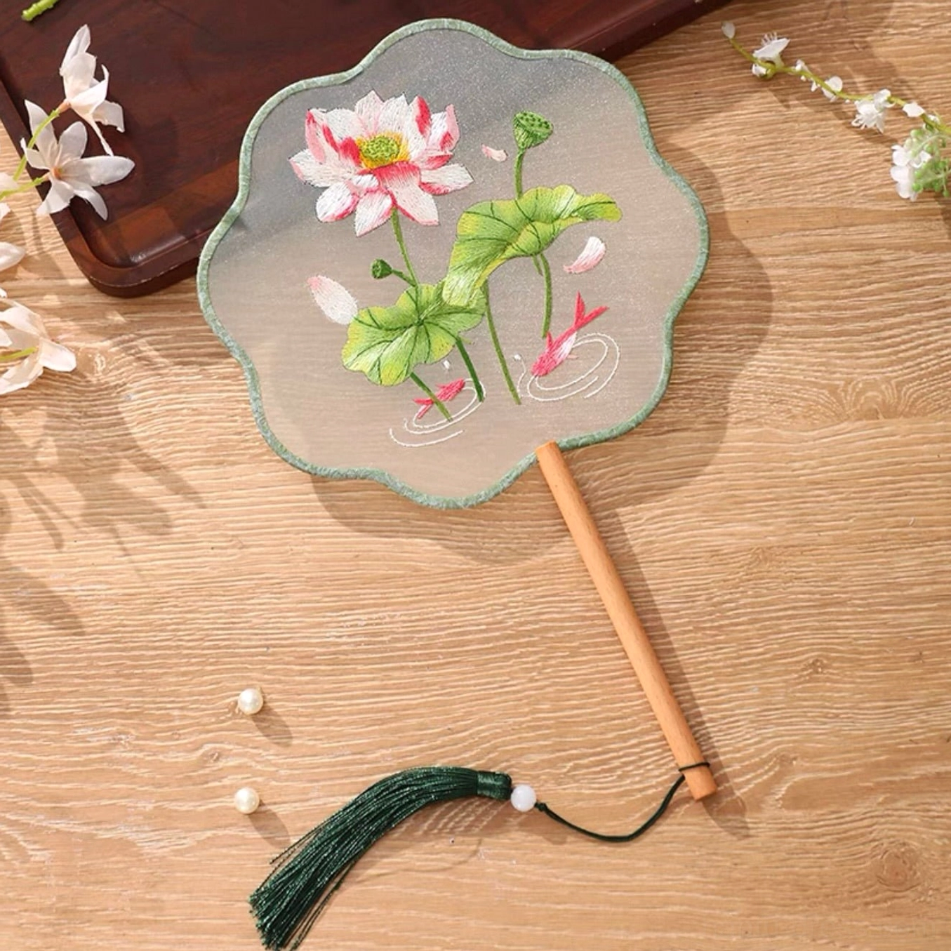Traditional Chinese tuanshan round hand fan with transparent embroidered lotus and koi design, light green petal edge, smooth wooden handle, and dark green tassel displayed on a warm wooden table