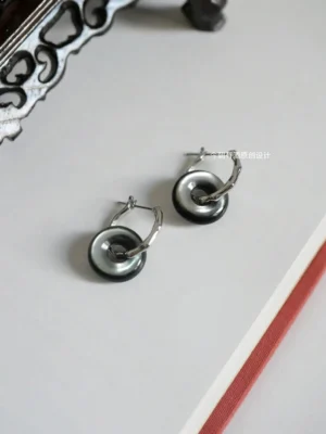 Lucky Loop Chinese earrings shown from an angled top view beside a carved Chinese ornament and red-trimmed book, highlighting the openable loop clasp and glossy gradient surface