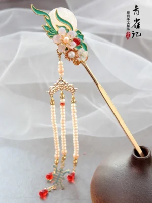 Side view of Luna Rain Chinese hair stick with pale moon ornament green cloisonne-style decoration floral beads and long red tassel beside a small teapot
