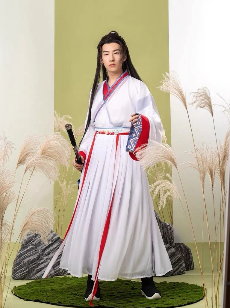 Male hanfu model wearing a white Wei Jin-inspired xianxia outfit with red and blue geometric collar and cuff trim, red sash, pleated skirt, and sword accessory