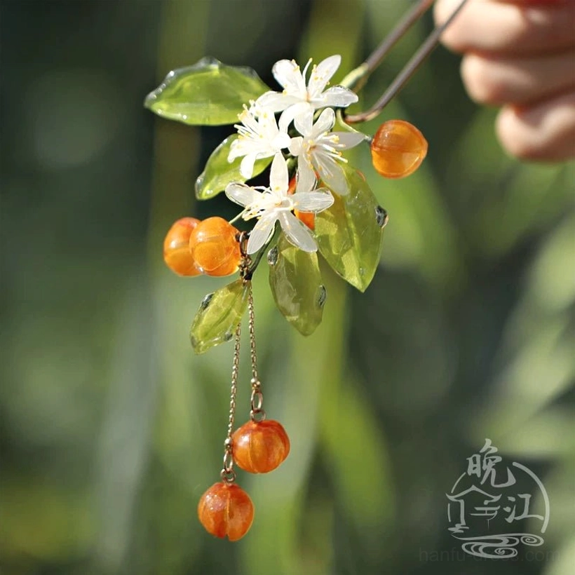 Handmade mandarin orange Chinese hair stick with green resin leaves white flowers amber orange fruit beads and dangling gold-tone pendants held against soft green foliage