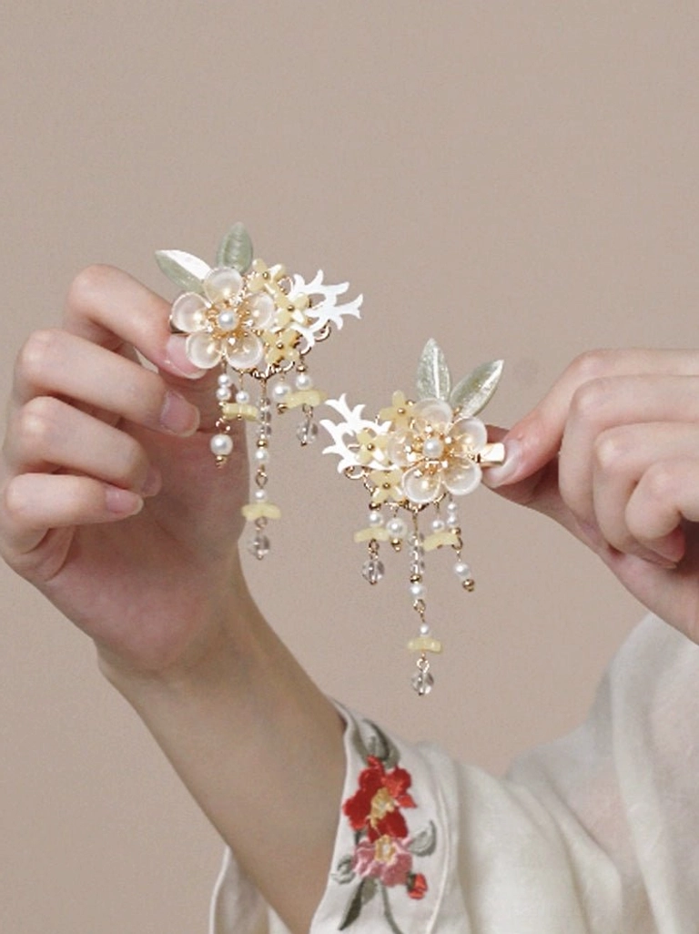 Matching pair of creamy yellow floral Hanfu hair clips with pearl and bead pendants held above embroidered traditional Chinese clothing cuffs
