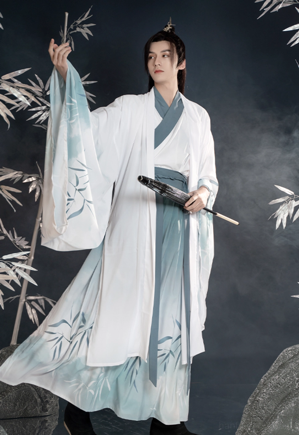 Three-quarter side view of a male model wearing Bamboo Rhyme men's Hanfu with turquoise waist sash, bamboo leaf print, flowing wide sleeves, folding fan, and bamboo-inspired wuxia styling