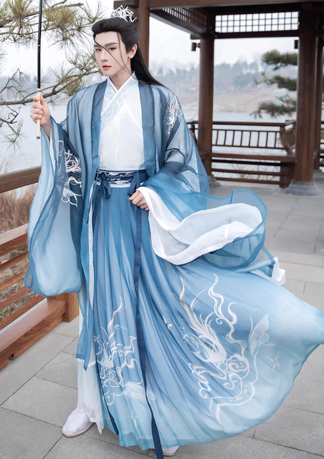 Blue gradient men's wuxia Hanfu outfit with large silver phoenix embroidery on the wrap skirt and white inner garment at a lakeside pavilion