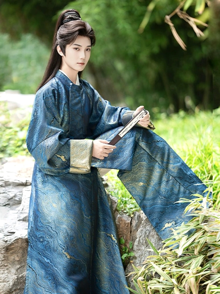 Three-quarter sitting view of a man in a deep blue Tang Dynasty Hanfu robe with gold-trimmed cuffs, flowing skirt-like hem, and folding fan in a garden