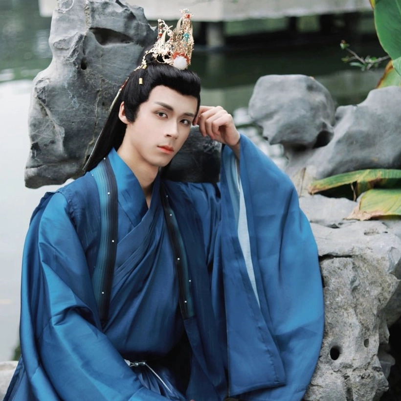 Male model wearing an elegant gold-tone Hanfu hair crown with red gemstone-style decorations and traditional blue Hanfu beside a waterside garden