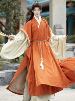 Male model wearing burnt orange Ming Dynasty winter Hanfu outer robe with cream fuzzy sleeve accents in a traditional Chinese stone courtyard