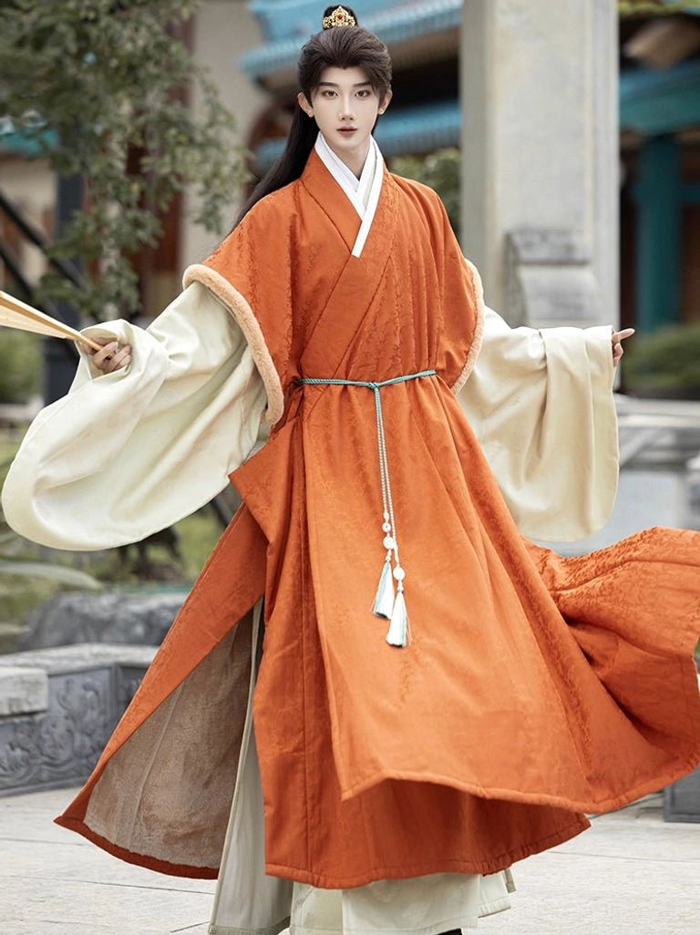 Male model wearing burnt orange Ming Dynasty winter Hanfu outer robe with cream fuzzy sleeve accents in a traditional Chinese stone courtyard