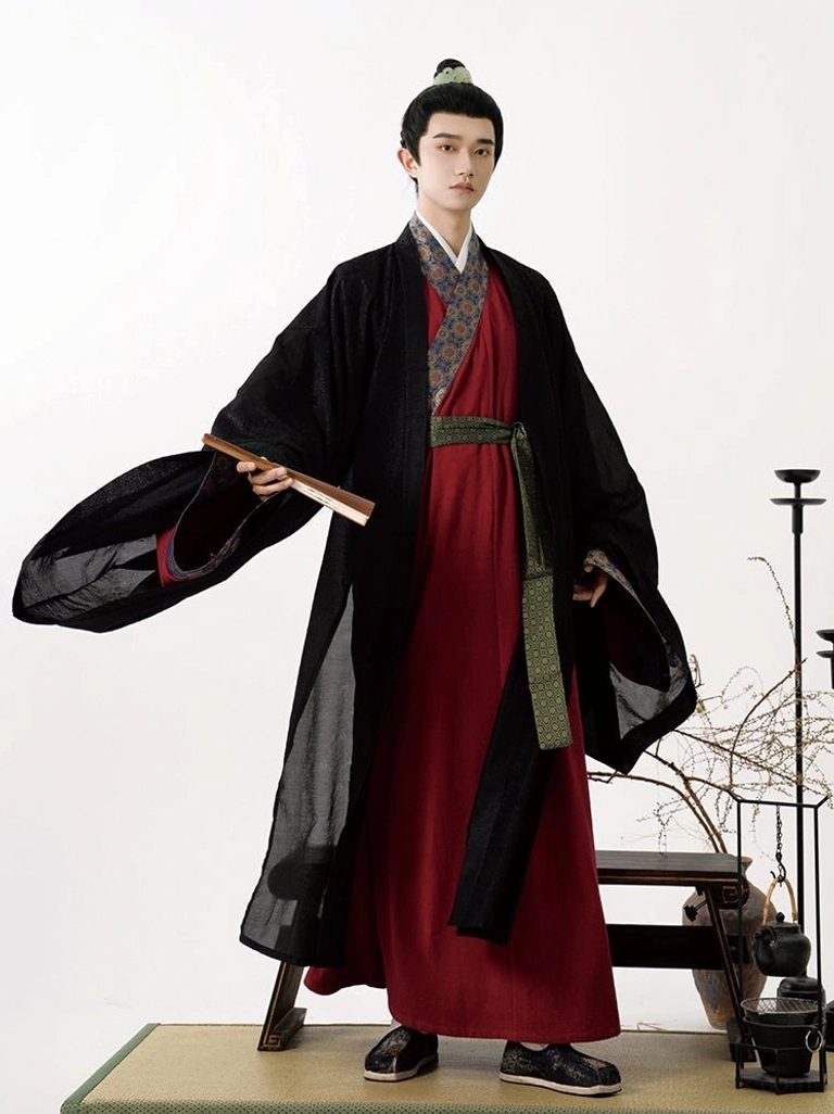 Three-quarter view of a man wearing traditional Song Dynasty-style hanfu with wide flared sleeves, patterned collar and cuffs, layered black robe, burgundy inner robe, and folding fan