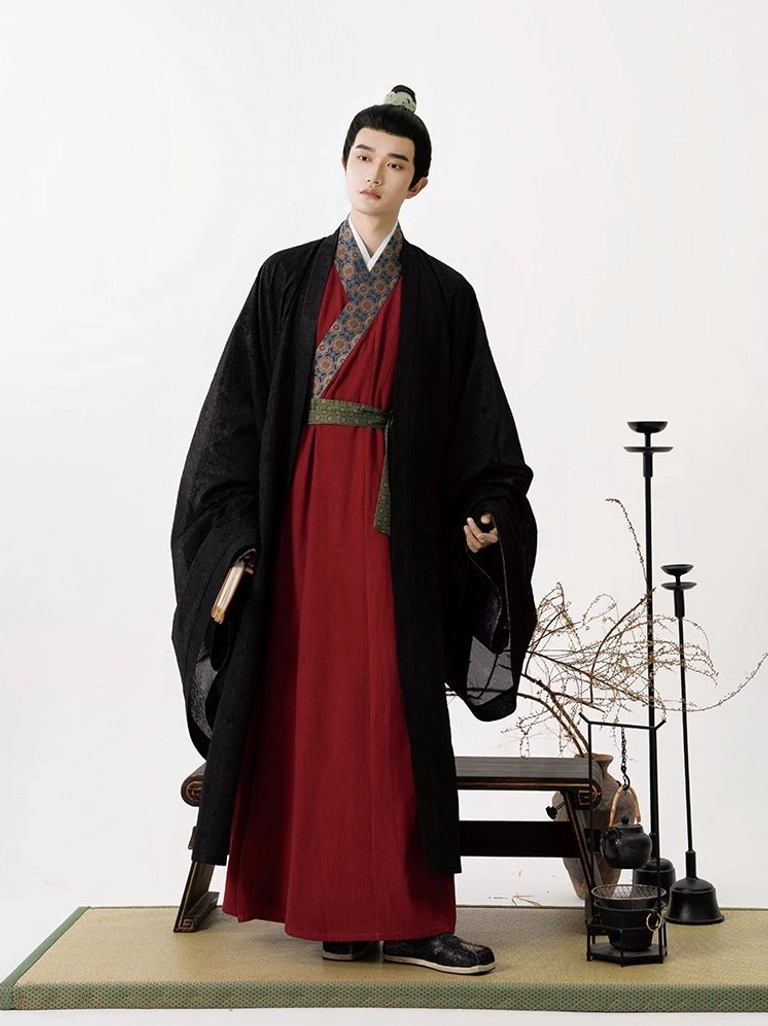 Male model wearing a Song Dynasty-inspired Jingchuan men’s hanfu outfit with a sheer black outer robe, burgundy red cross-collar robe, wide flowing sleeves, patterned trim, and dark green waist sash