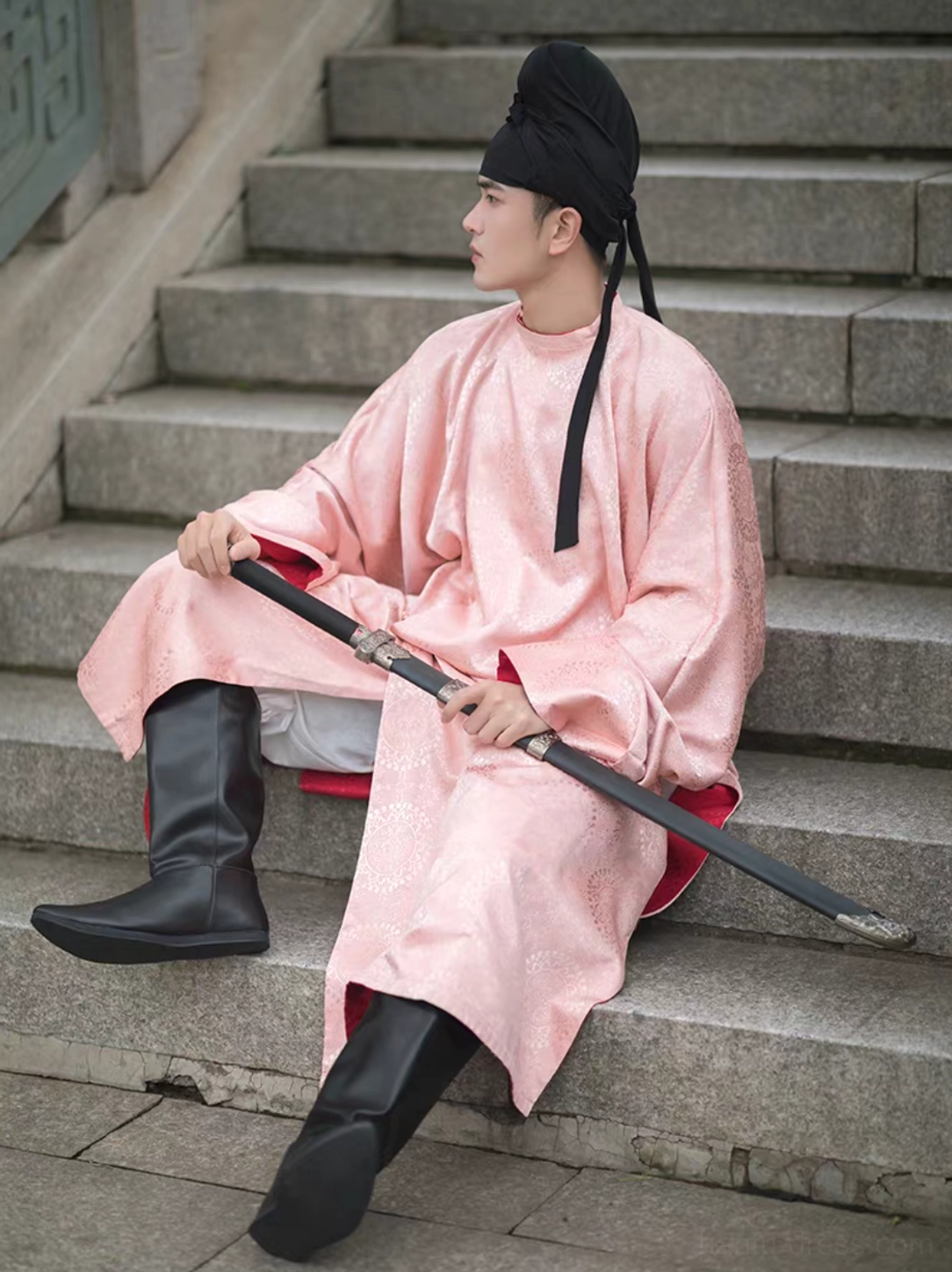 Full-body male model wearing a pink Tang-style Hanfu outfit with black high-tube pointed Hanfu boots while seated on ancient stone stairs with a traditional sword