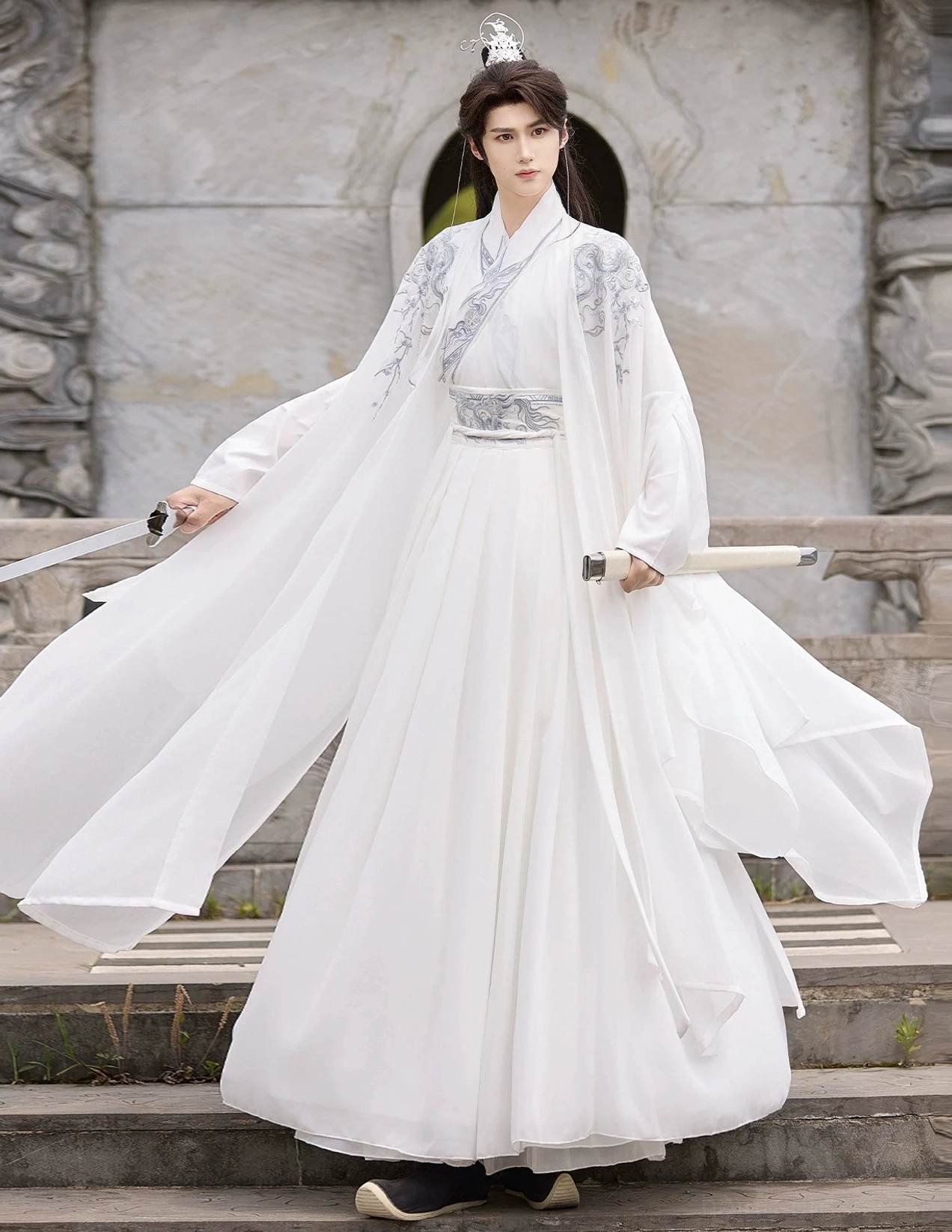 Full-body male model wearing an all-white men's Wei-Jin Hanfu cross-collar knight outfit with silver shoulder and waist patterns, flowing sheer outer coat, pleated wrap skirt, traditional Chinese hair crown, and sword props on outdoor stone stairs