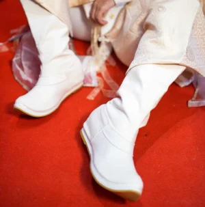 White men's traditional Chinese Hanfu high-top boots with upturned toes and tan rubber soles worn with a light beige patterned Hanfu outfit on a red carpet