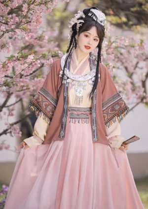 Miao-inspired Hanfu costume set shown from the front beneath pink cherry blossoms, featuring ornate embroidered jacket cuffs and hem, a high-waisted pink skirt, and a traditional-inspired silver pendant
