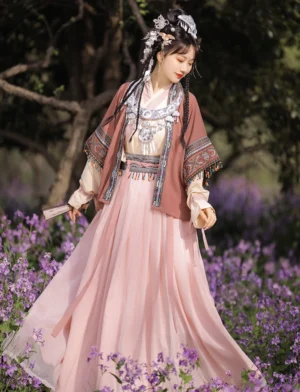 Miao-inspired modern Hanfu three-piece costume set shown from a full-body side view in a flower garden, featuring an embroidered warm-toned jacket, soft pink flowing skirt, silver-inspired accessories, and braided hair ornaments