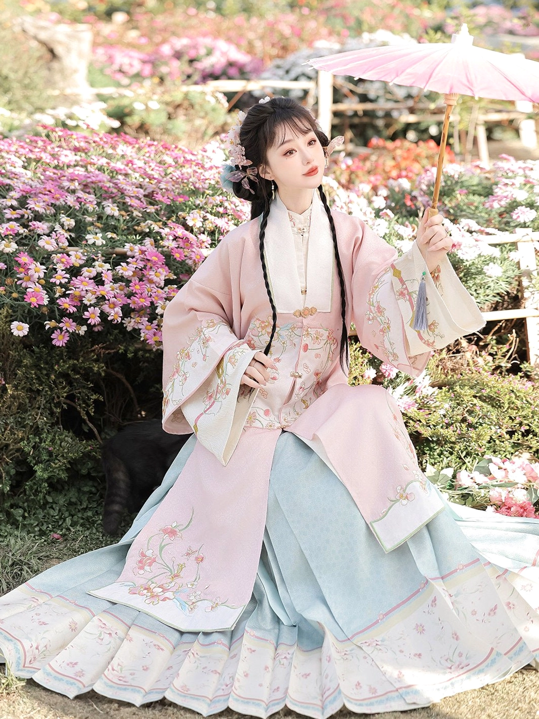 Pink embroidered Ming Dynasty Hanfu robe and soft blue mamian skirt shown seated among flowering garden shrubs with pink umbrella
