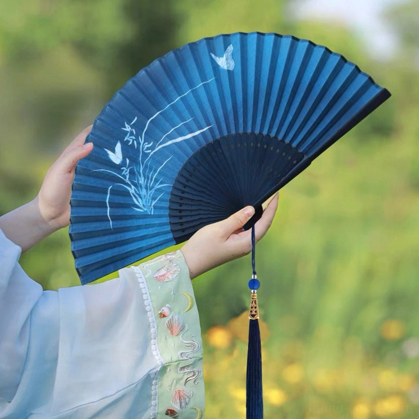 Midnight blue Chinese folding hand fan with white orchid and butterfly artwork, black bamboo ribs, and dark blue tassel held with embroidered Hanfu sleeves in a flower field