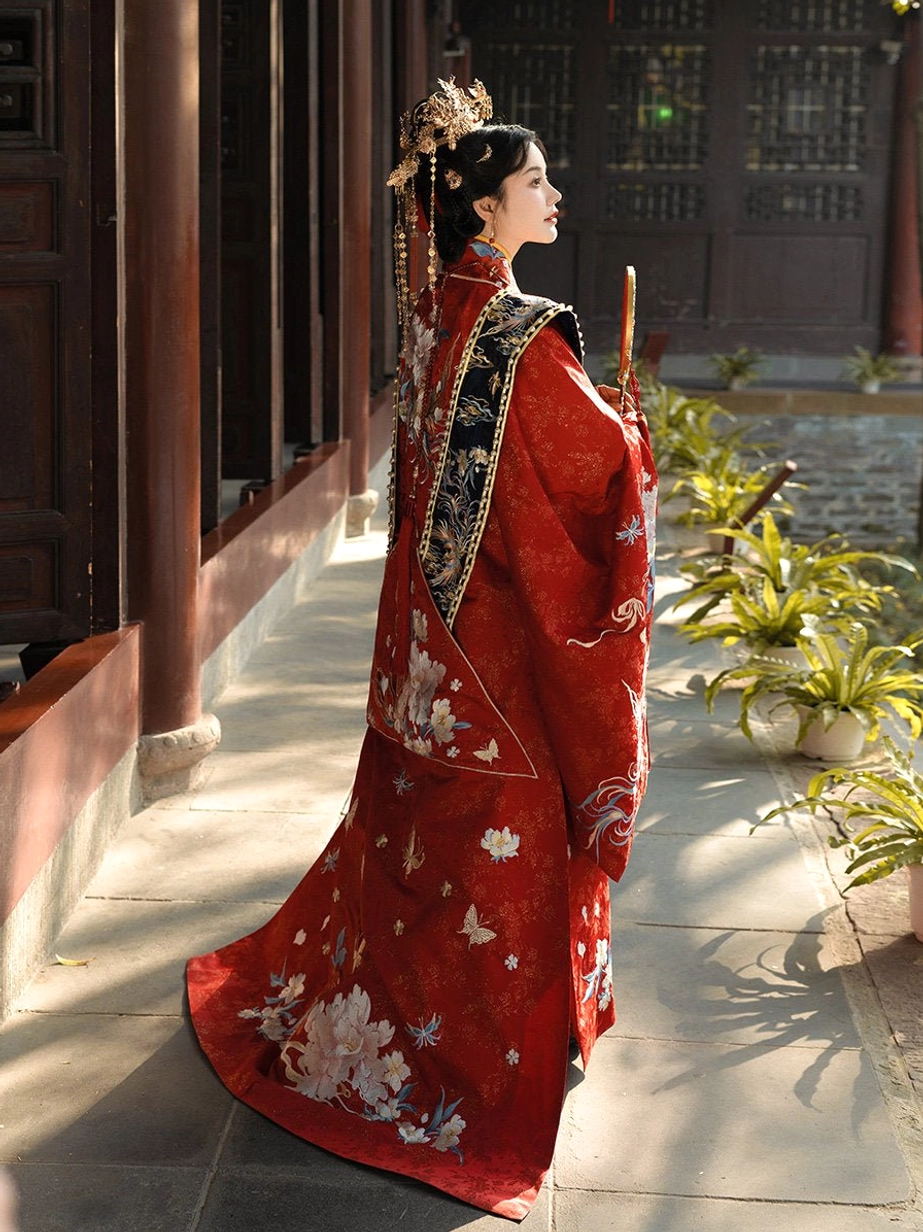 Side rear view of a bride wearing scarlet Ming Dynasty Chinese wedding Hanfu with peony and butterfly embroidery, black and gold back sash, traditional hair ornaments, and flowing bridal train